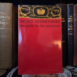 May include: A red book titled "MOSES MAIMONIDES the guide for the perplexed" with gold and black accents. The book is surrounded by other antique books. The cover has Hebrew text and a crown illustration.