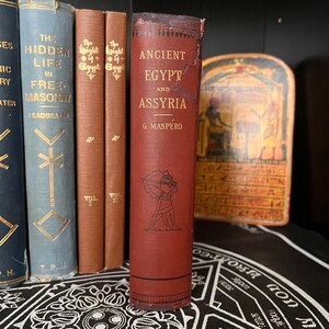以下が含まれることがあります： G. マスペロ著「古代エジプトとアッシリア」という赤いハードカバーの本。本は装飾的な模様のある黒い表面の上に置かれています。