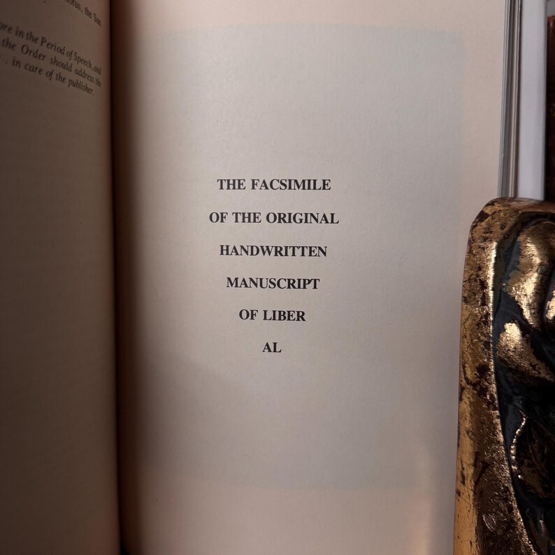 May include: An open book page with text that reads "THE FACSIMILE OF THE ORIGINAL HANDWRITTEN MANUSCRIPT OF LIBER AL." The text is in a serif font and is centered on the page. The book's pages are off-white, and the text is black.