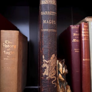 Puede incluir: Una colecci&oacute;n de libros antiguos en una estanter&iacute;a. La columna vertebral del libro "Barrett's Magus" es visible, con letras doradas y una ilustraci&oacute;n de una figura con un drag&oacute;n. Otros libros incluyen "The History of Magic".