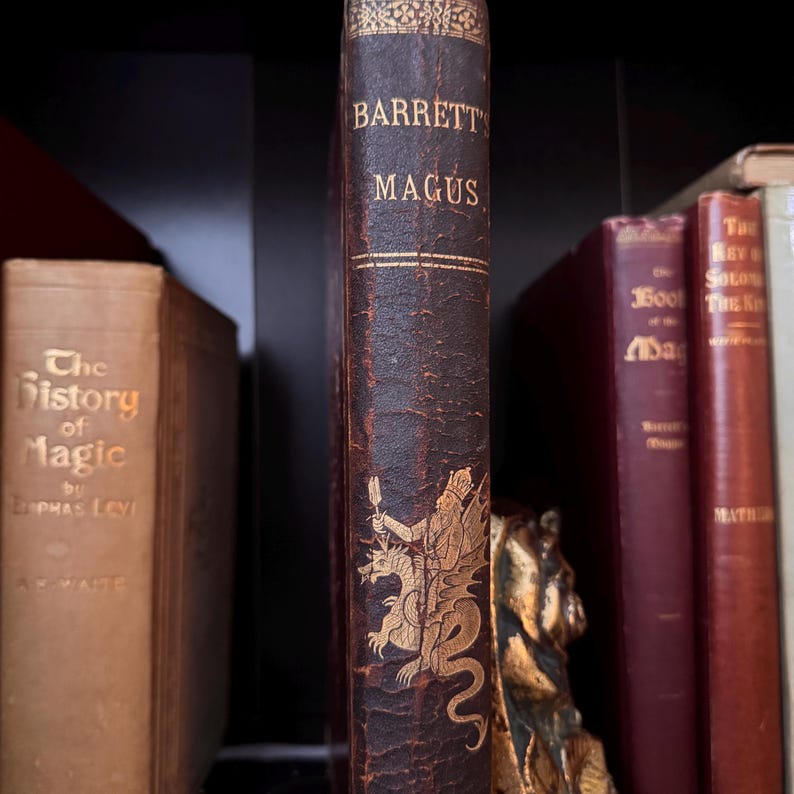 Puede incluir: Una colecci&oacute;n de libros antiguos en una estanter&iacute;a. La columna vertebral del libro "Barrett's Magus" es visible, con letras doradas y una ilustraci&oacute;n de una figura con un drag&oacute;n. Otros libros incluyen "The History of Magic".