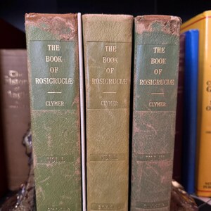 Może przedstawiać: Trzy stare zielone książki z pozłacanymi napisami zatytułowane "The Book of Rosicruciae" autorstwa Clymera. Książki to tomy I, II i III.