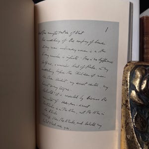 May include: An open book with handwritten text on a light gray page. The text is in black ink and appears to be a poem or a passage from a book. A decorative gold and black object is visible in the background.
