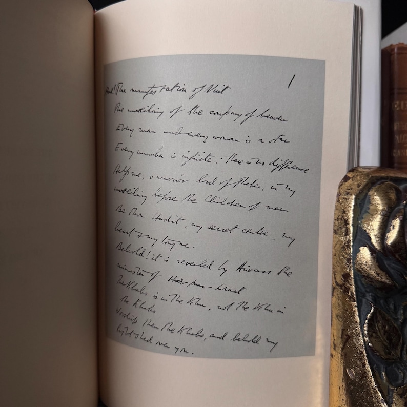 May include: An open book with handwritten text on a light gray page. The text is in black ink and appears to be a poem or a passage from a book. A decorative gold and black object is visible in the background.