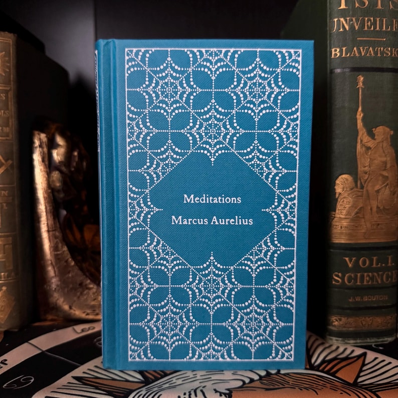 May include: A teal-colored hardcover book titled "Meditations" by Marcus Aurelius. The cover features a white, intricate web-like design. Other antique books are visible in the background, suggesting a collection of classic literature.