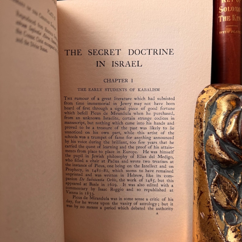 May include: An open book displaying the title "THE SECRET DOCTRINE IN ISRAEL." The text discusses Kabalism and historical figures. The book has a decorative spine, and the pages are aged.