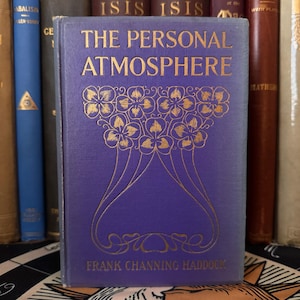 Puede incluir: Un libro antiguo titulado "The Personal Atmosphere" con letras doradas y un dise&ntilde;o floral en una cubierta morada. El nombre del autor, Frank Channing Haddock, est&aacute; impreso en la parte inferior. El libro est&aacute; de pie entre otros libros.