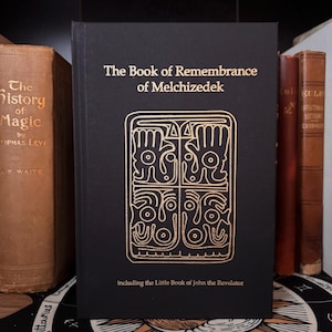 Peut inclure: Un livre noir intitulé "The Book of Remembrance of Melchizedek" avec des lettres dorées et un motif géométrique doré. Le livre comprend "The Little Book of John the Revelator". D'autres livres sont visibles en arrière-plan.