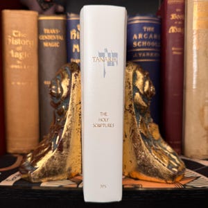 May include: A white book titled "Tanakh: The Holy Scriptures" with gold lettering, standing between two ornate gold bookends. Other books with gold lettering and dark covers are visible in the background.