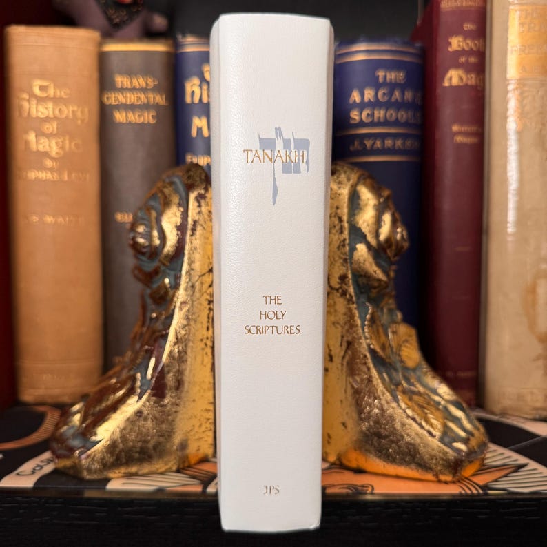 May include: A white book titled "Tanakh: The Holy Scriptures" with gold lettering, standing between two ornate gold bookends. Other books with gold lettering and dark covers are visible in the background.