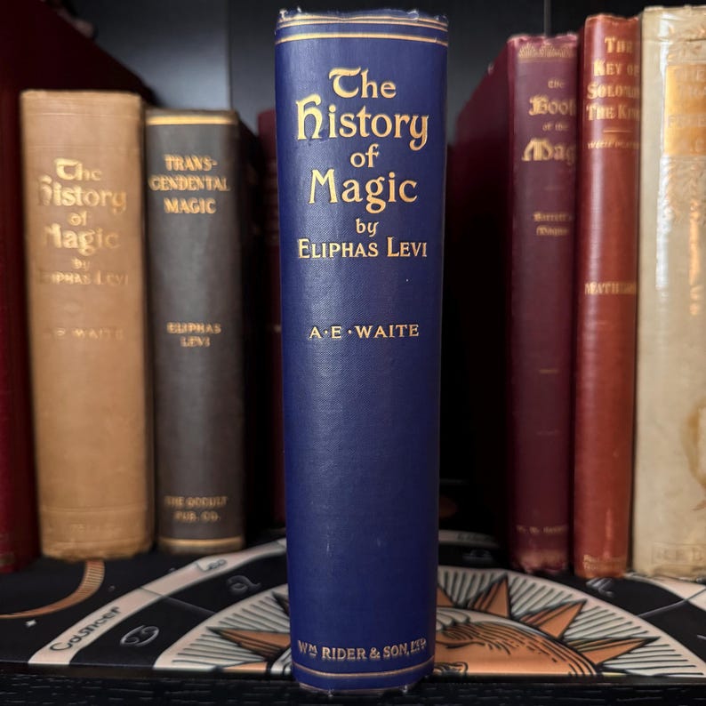 Puede incluir: Un libro de tapa dura azul titulado "The History of Magic" de Eliphas Levi, con el nombre del autor y "A.E. Waite" impresos en letras doradas. El libro est&aacute; en un estante con otros libros.