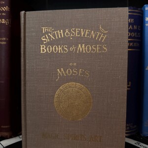 May include: A brown book with gold lettering on the cover. The title is "The Sixth & Seventh Books of Moses or Moses Magic, Spirits, Art."