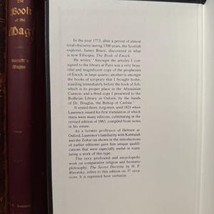 May include: A close-up of a book page with text from "The Book of Enoch" and "The Secret Doctrine" by H.P. Blavatsky. The text is in black font on a cream-colored page. The book is next to other books with dark burgundy covers.