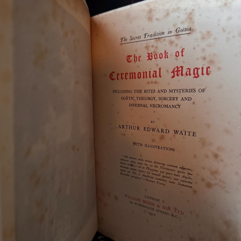 May include: Open book titled "The Book of Ceremonial Magic" by Arthur Edward Waite. The cover is a light tan color with red and black text. The book includes illustrations and was published in London in 1911.