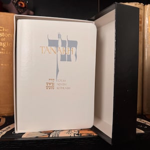 May include: A white book with the word "TANAKH" in gold lettering and Hebrew text below. The book is in a black box, with other books in the background.
