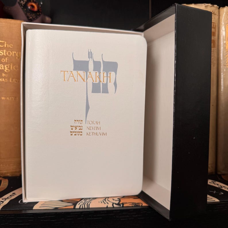 May include: A white book with the word "TANAKH" in gold lettering and Hebrew text below. The book is in a black box, with other books in the background.