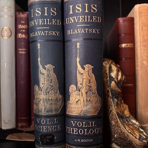 May include: Two antique books titled "Isis Unveiled" by Blavatsky. The dark blue books have gold lettering and illustrations of a robed figure. Volume I is labeled "Science" and Volume II is labeled "Theology". Other books are visible on the shelf.