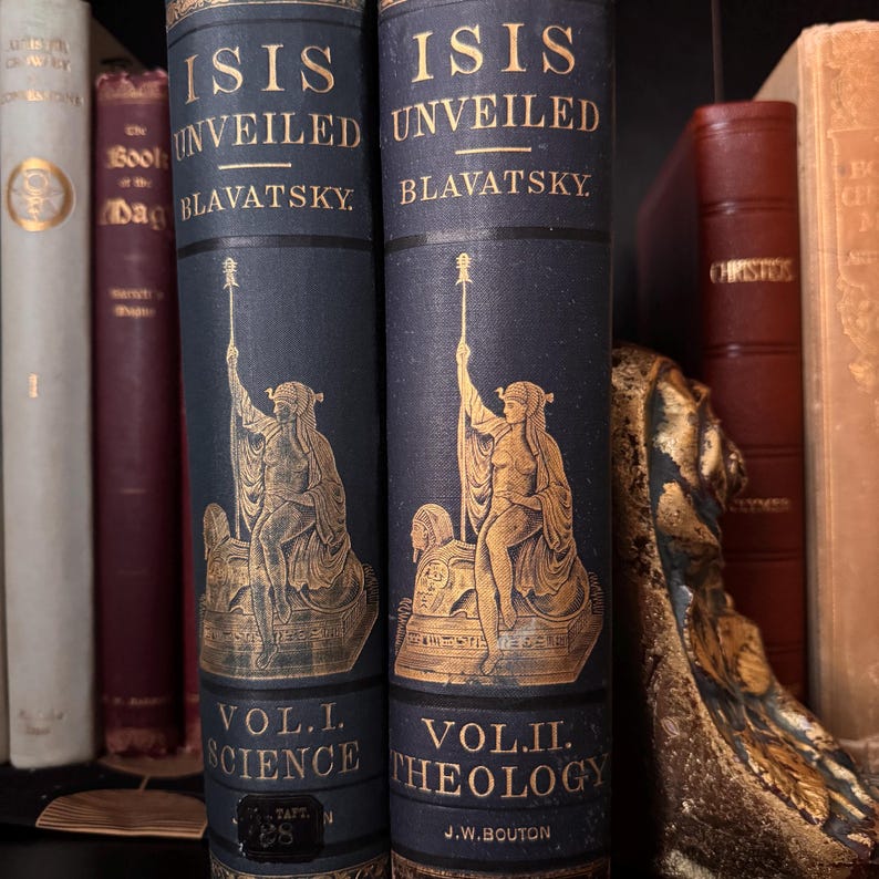 May include: Two antique books titled "Isis Unveiled" by Blavatsky. The dark blue books have gold lettering and illustrations of a robed figure. Volume I is labeled "Science" and Volume II is labeled "Theology". Other books are visible on the shelf.