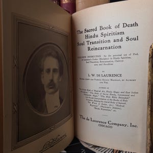May include: Open book with the title "The Sacred Book of Death Hindu Spiritism Soul Transition and Soul Reincarnation." The book's pages are aged, with text and a portrait of a man in a frame. The author is L.W. De Laurence.