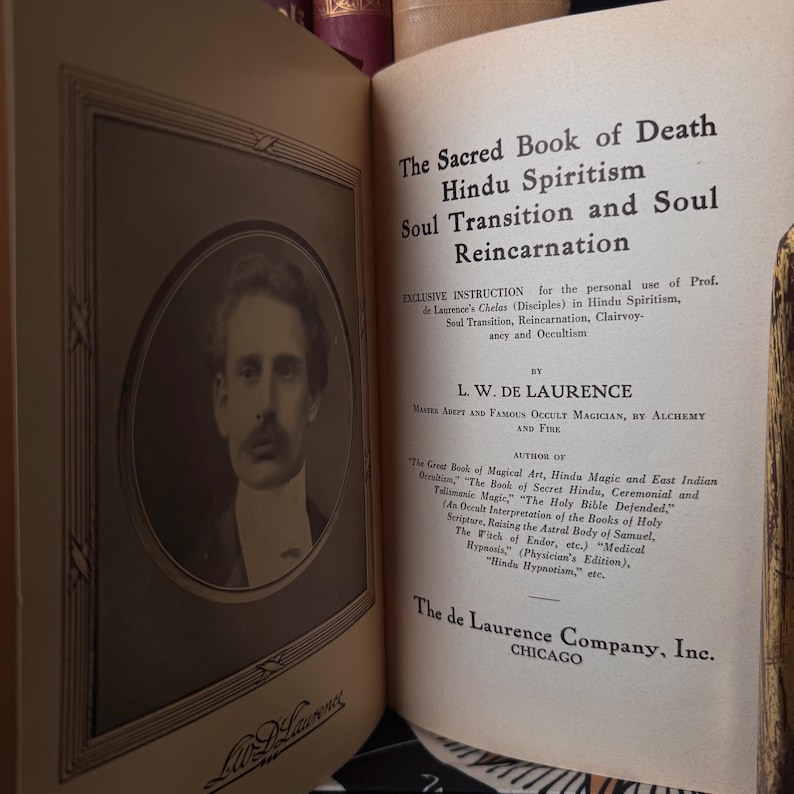 May include: Open book with the title "The Sacred Book of Death Hindu Spiritism Soul Transition and Soul Reincarnation." The book's pages are aged, with text and a portrait of a man in a frame. The author is L.W. De Laurence.