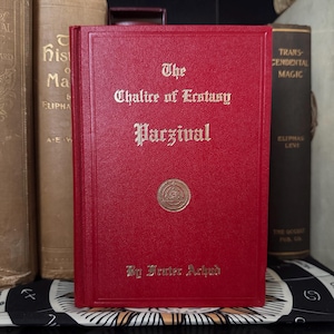 Könnte beinhalten: Ein rotes Hardcover-Buch mit dem Titel "The Chalice of Ecstasy Parzival" mit goldener Schrift und einem goldenen Emblem. Das Buch ist von Frater Achad und Teil einer Sammlung okkulter Bücher.