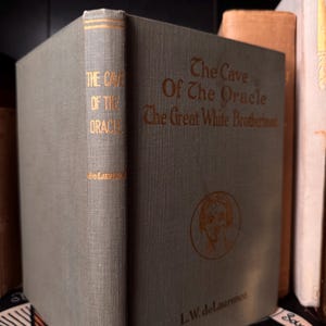 May include: A vintage book titled "The Cave Of The Oracle" with gold lettering on a gray-green cover. The spine reads "The Cave Of The Oracle" vertically. The book is part of a collection, with other books visible.