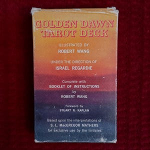 Könnte beinhalten: Eine Schachtel für ein Golden Dawn Tarot Deck, illustriert von Robert Wang. Die Schachtel ist orange, gelb und rot mit schwarzem Text. Der Text lautet "Golden Dawn Tarot Deck", "Illustriert von Robert Wang", "Unter der Leitung von Israel Regardie", "Komplett mit Booklet mit Anweisungen von Robert Wang", "Vorwort von Stuart R. Kaplan", "Basierend auf den Interpretationen von S. L. MacGregor Mathers für den exklusiven Gebrauch durch die Eingeweihten".