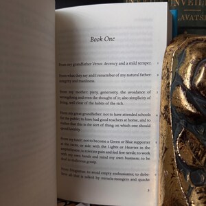 May include: An open book with the title "Book One" visible on the page. The text includes numbered paragraphs. A decorative gold-colored object is partially visible on the right side of the image, with a blurred background.
