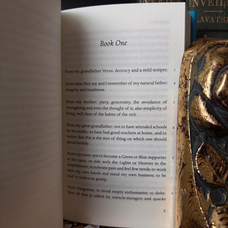 May include: An open book with the title "Book One" visible on the page. The text includes numbered paragraphs. A decorative gold-colored object is partially visible on the right side of the image, with a blurred background.