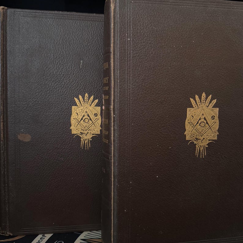 Puede incluir: Dos libros antiguos encuadernados en cuero marr&oacute;n. Las cubiertas presentan un emblema dorado con un tri&aacute;ngulo, un ojo y otros s&iacute;mbolos. Los libros probablemente contengan textos hist&oacute;ricos o religiosos, con letras doradas en los lomos.