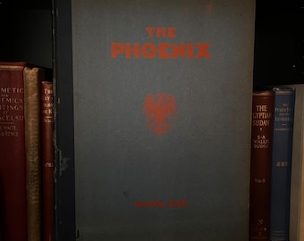 Może przedstawiać: Antyczna książka w twardej oprawie zatytułowana "The Phoenix" autorstwa Manly'ego Halla. Okładka książki jest ciemno szaro-niebieska z czerwonym wzorem ptaka feniksa w środku. Książka znajduje się na półce z innymi książkami.