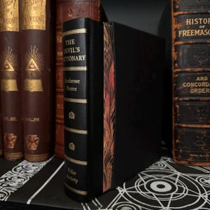 May include: A black leather-bound book titled "The Devil's Dictionary" by Ambrose Bierce. The book is part of the Folio Society collection and has a decorative gold spine with a marbled design.
