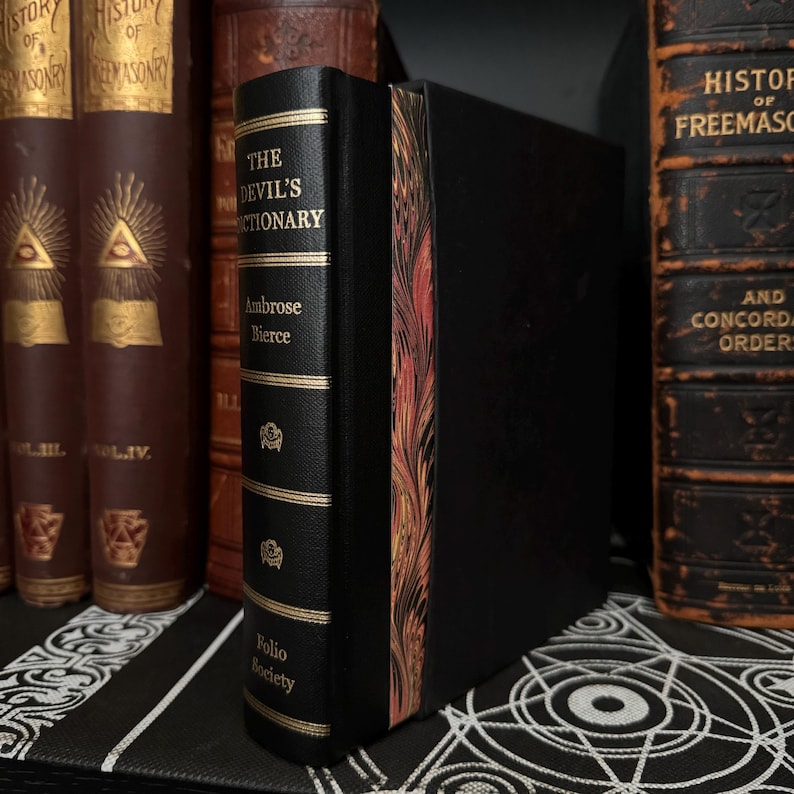 May include: A black leather-bound book titled "The Devil's Dictionary" by Ambrose Bierce. The book is part of the Folio Society collection and has a decorative gold spine with a marbled design.