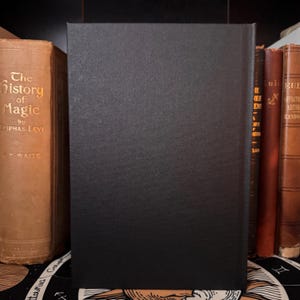 Peut inclure: Un livre &agrave; couverture noire et textur&eacute;e est pr&eacute;sent&eacute; debout, entour&eacute; d'autres livres anciens. Le titre "The History of Magic" est visible sur un livre &agrave; gauche. L'ensemble &eacute;voque un th&egrave;me de myst&egrave;re et d'histoire.