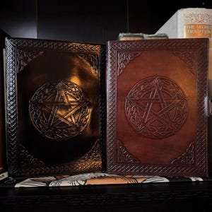May include: Two leather-bound books with embossed pentagram designs. One book is black, the other is brown. Both have Celtic knotwork borders and a central pentagram within a circular frame. The books are displayed on a dark surface.
