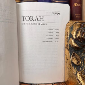 May include: An open book with the title "TORAH" and the subtitle "THE FIVE BOOKS OF MOSES" is displayed. The names of the books of the Torah are listed below the title. The book is open to a white page with black text. The book is surrounded by other books.