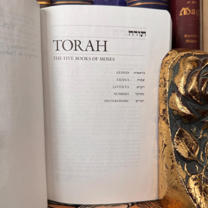 May include: An open book with the title "TORAH" and the subtitle "THE FIVE BOOKS OF MOSES" is displayed. The names of the books of the Torah are listed below the title. The book is open to a white page with black text. The book is surrounded by other books.
