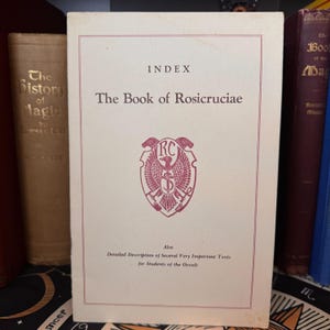Może przedstawiać: Biała książka z tytułem "The Book of Rosicruciae" i słowami "Index" oraz "Also Detailed Description of Several Very Important Texts for Students of the Occult". Książka ma czerwony i biały emblemat z literami "RC" w środku.