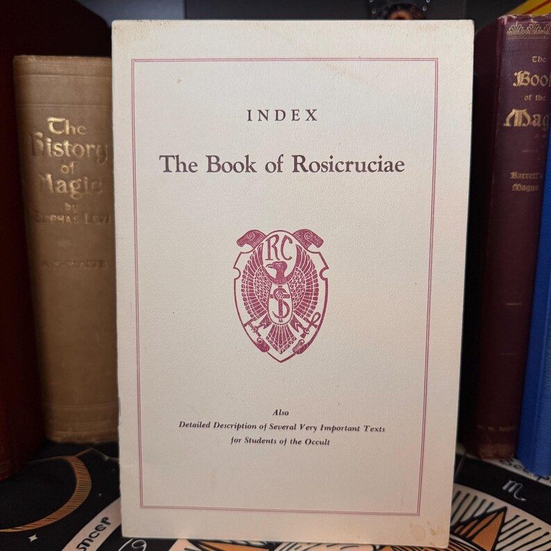 Może przedstawiać: Biała książka z tytułem "The Book of Rosicruciae" i słowami "Index" oraz "Also Detailed Description of Several Very Important Texts for Students of the Occult". Książka ma czerwony i biały emblemat z literami "RC" w środku.