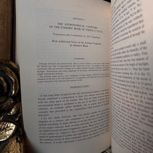 May include: Open book with text titled "THE ASTRONOMICAL CHAPTERS OF THE ETHIOPIC BOOK OF ENOCH." The pages are off-white with black text. The book is open to a section with a summary and introduction. The book is on a wooden surface.