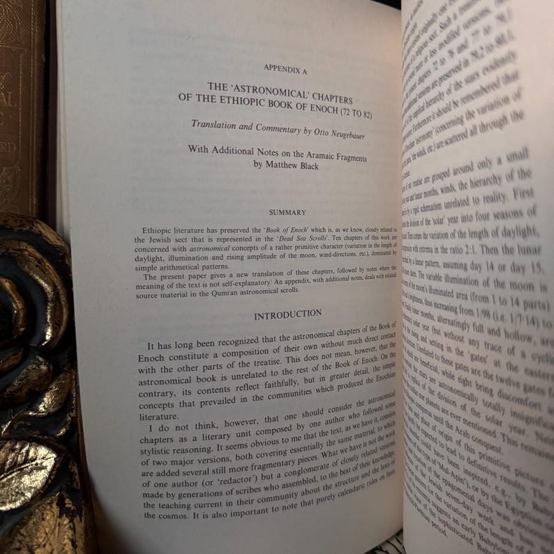 May include: Open book with text titled "THE ASTRONOMICAL CHAPTERS OF THE ETHIOPIC BOOK OF ENOCH." The pages are off-white with black text. The book is open to a section with a summary and introduction. The book is on a wooden surface.