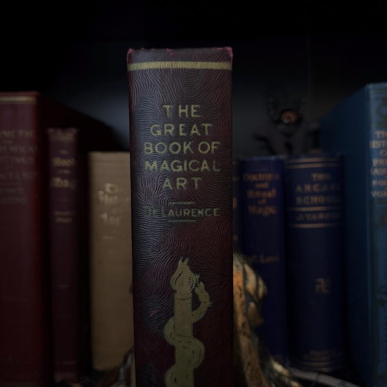 Pu&ograve; includere: Un libro vintage intitolato "The Great Book of Magical Art" di De Laurence. Il libro ha una copertina color borgogna con un titolo in rilievo dorato e un'illustrazione stilizzata di un mago con una bacchetta.