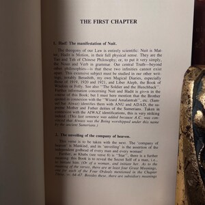 May include: An open book page with the title "THE FIRST CHAPTER" at the top. The text discusses the manifestation of Nuit, the theogony of Law, and the unveiling of the company of heaven. The text is in black font.
