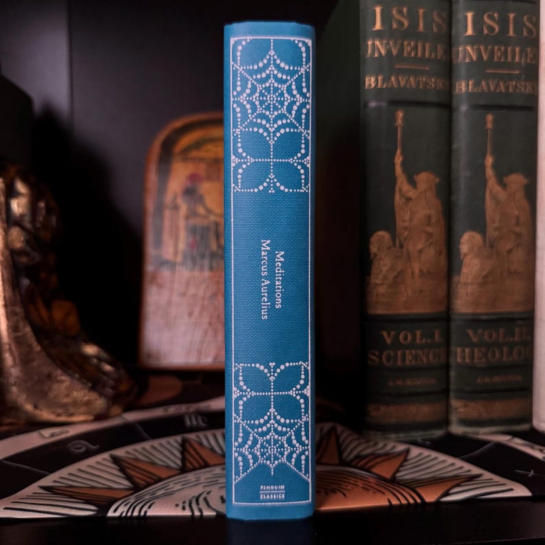May include: A teal-colored book titled "Meditations" by Marcus Aurelius. The spine features white decorative patterns and text. Other books with gold lettering are visible in the background, along with a decorative shelf.