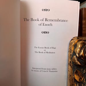 May include: Open book with the title "The Book of Remembrance of Enoch" printed in black on a white page. Below the title are the words "The Essene Book of Hagi or The Book of Meditation". The text "Interpreted from stone tablets by means of Urim & Thummim" is also visible.