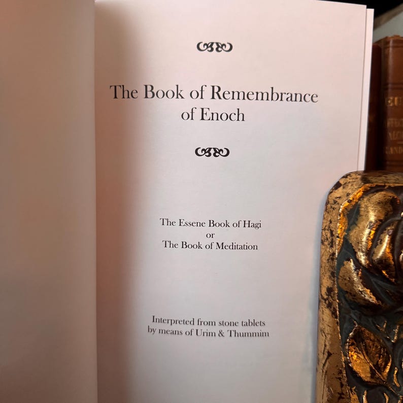 May include: Open book with the title "The Book of Remembrance of Enoch" printed in black on a white page. Below the title are the words "The Essene Book of Hagi or The Book of Meditation". The text "Interpreted from stone tablets by means of Urim & Thummim" is also visible.