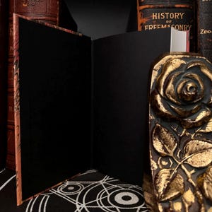 May include: An open, black, blank journal with a marbled cover. The journal is resting on a black and white patterned surface. A gold rose bookend is visible on the right side of the image.