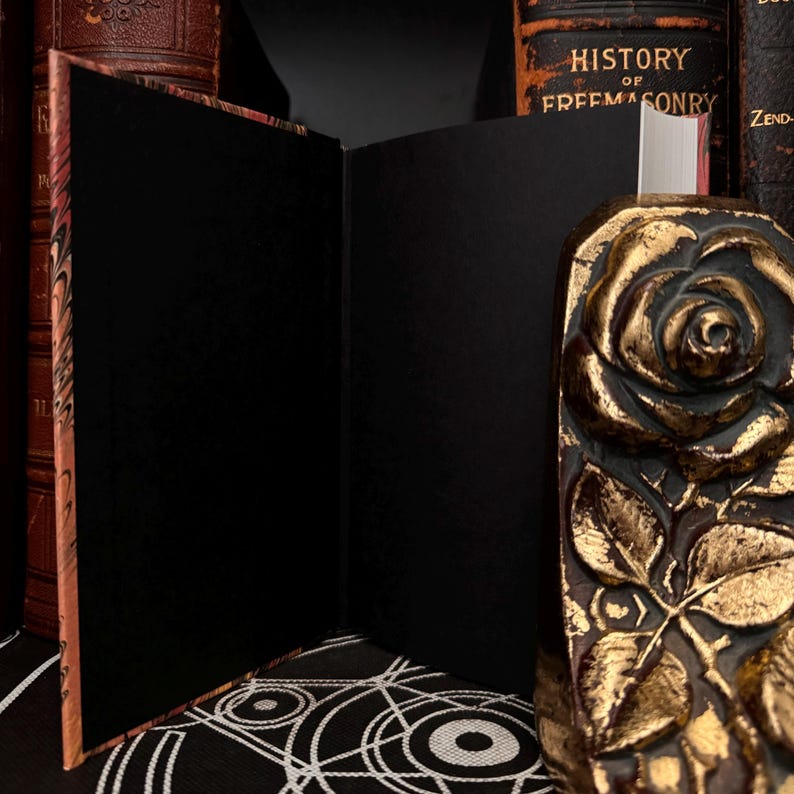 May include: An open, black, blank journal with a marbled cover. The journal is resting on a black and white patterned surface. A gold rose bookend is visible on the right side of the image.