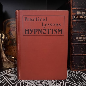 May include: A red hardcover book with the title "Practical Lessons in Hypnotism" printed in black.