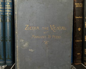 May include: A blue hardcover book with gold lettering on the spine and front cover. The title is "Zenia the Vestal" by Margaret B. Peeke.
